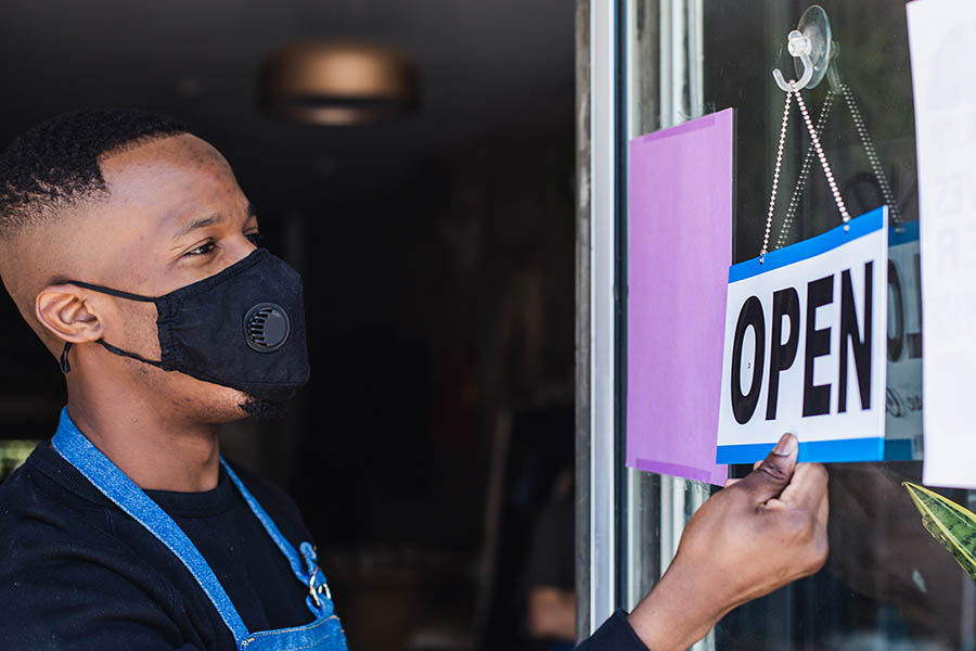Man turning shop sign from 'Closed' to 'Open' while wearing a face mask